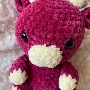 Könnte beinhalten: Ein gehäkeltes Plüschtier in Form einer Kuh. Die Kuh ist rosa mit weißen Akzenten an den Hörnern, der Nase und den Füßen. Die Kuh hat schwarze Augen und einen süßen Ausdruck.
