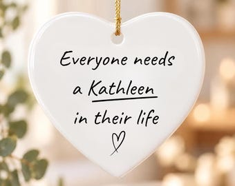 Puede incluir: Un adorno de cerámica blanco en forma de corazón que cuelga de un cordón dorado. El texto "Everyone needs a Kathleen in their life" está impreso en negro, con el nombre subrayado y un pequeño corazón dibujado debajo.