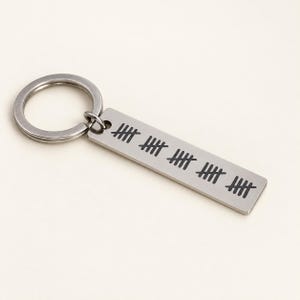 Puede incluir: Un llavero rectangular de metal plateado con una anilla unida a una placa plana. La superficie de la placa est&aacute; grabada con cinco grupos de marcas de conteo en color negro, cada una formada por cuatro l&iacute;neas verticales cruzadas por una diagonal.
