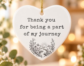 Puede incluir: Un adorno de cerámica blanco en forma de corazón que cuelga de un cordón dorado. El texto "Thank you for being a part of my journey" está impreso en negro, junto a un dibujo lineal negro de un camino sinuoso, flores y un sol naciente.