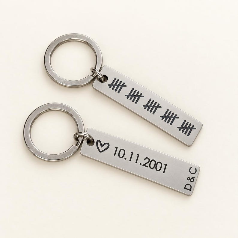 Puede incluir: Dos llaveros rectangulares de metal plateado con anillas. Un llavero presenta cinco conjuntos de marcas de conteo. El otro llavero muestra un s&iacute;mbolo de coraz&oacute;n, la fecha 10.11.2001 y las letras D&C grabadas en negro sobre la superficie.