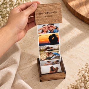 May include: A square wooden box with a hinged lid featuring the engraved text Henry Amelia 10/10/2021. The lid is lifted to reveal a vertical accordion-style strip of five color photographs depicting a couple in various outdoor settings.