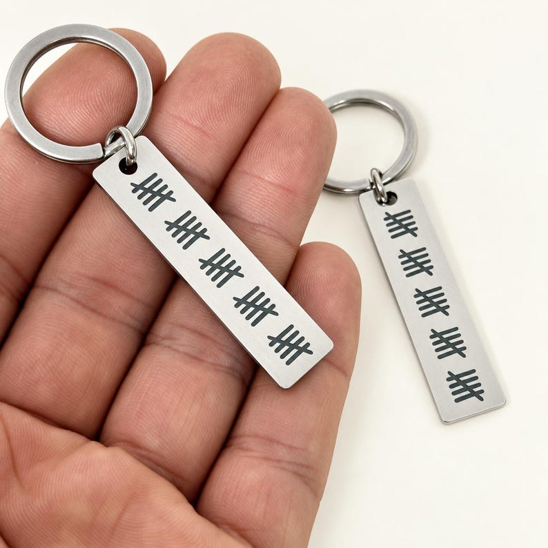 Puede incluir: Dos llaveros rectangulares de metal plateado con una anilla en la parte superior. Cada llavero est&aacute; grabado con una serie de marcas de conteo negras dispuestas en grupos de cinco, representando un total de veinticinco por pieza.