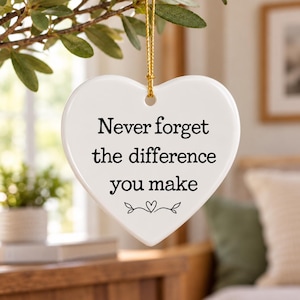 以下が含まれることがあります： Never forget the difference you makeという黒い文字と、その下に小さなハートと葉のグラフィックがプリントされた、白いハート型のセラミック製オーナメント。細い金の紐で吊るされており、背景はぼやけています。
