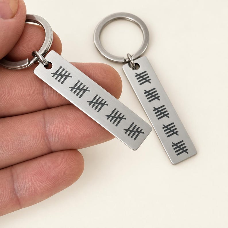 Puede incluir: Dos llaveros rectangulares de metal plateado con dise&ntilde;os de marcas de conteo grabadas. Cada llavero est&aacute; unido a una argolla met&aacute;lica circular. Las marcas est&aacute;n dispuestas en grupos de cinco, con cuatro l&iacute;neas verticales cruzadas por una diagonal.