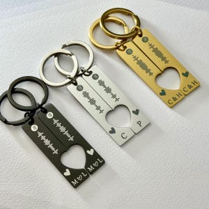 May include: Three keyrings with a heart-shaped cutout, each split between two rectangular tags. One is black, one silver, and one gold. Each tag has a Spotify code and initials. The gold keyring has the initials "C & H". The silver keyring has the initials "C & P". The black keyring has the initials "M & L".