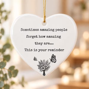 May include: A white heart-shaped ceramic ornament featuring black text that reads: Sometimes amazing people forget how amazing they are... This is your reminder. Below the text is a black line drawing of a flower bouquet, a bee, and a butterfly.