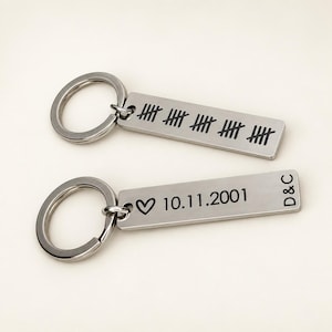 Puede incluir: Dos llaveros rectangulares de metal plateado con anillas. Uno muestra cuatro grupos de cinco marcas de conteo. El otro presenta un s&iacute;mbolo de coraz&oacute;n, la fecha 10.11.2001 y las letras D&C grabadas en color negro.