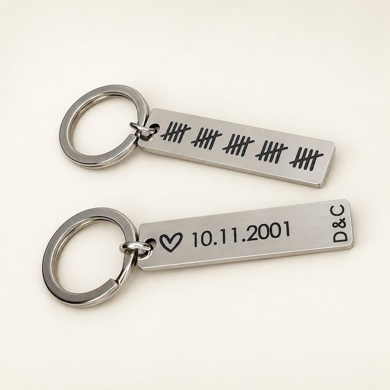 Puede incluir: Dos llaveros rectangulares de metal plateado con anillas. Uno muestra cuatro grupos de cinco marcas de conteo. El otro presenta un s&iacute;mbolo de coraz&oacute;n, la fecha 10.11.2001 y las letras D&C grabadas en color negro.