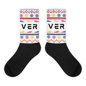 Puede incluir: Par de calcetines blancos y negros con un diseño festivo. Los calcetines presentan la palabra "VER" en negro, con un patrón de copos de nieve, coches y formas geométricas en rojo, amarillo y azul. La parte superior de los calcetines tiene un borde rojo y azul.