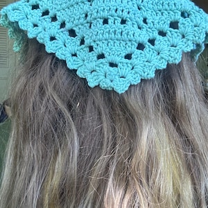 May include: A turquoise crochet head covering with a scalloped edge and openwork design. The head covering is worn over long, wavy, light-colored hair. The crochet pattern features small, open holes.