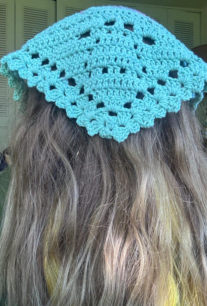 May include: A turquoise crochet head covering with a scalloped edge and openwork design. The head covering is worn over long, wavy, light-colored hair. The crochet pattern features small, open holes.