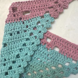 May include: A handmade crochet shawl in a triangular shape. The shawl features a two-tone design with alternating sections of dusty rose and light turquoise. The openwork pattern adds a delicate touch to the soft, textured yarn.