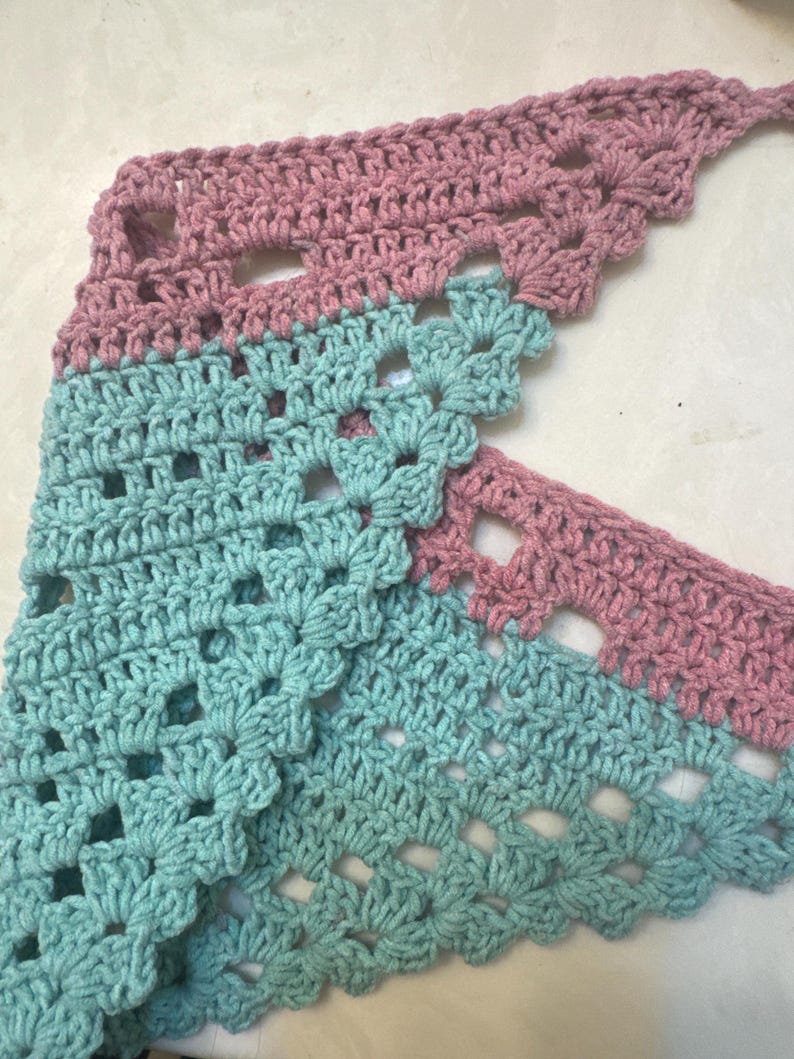 May include: A handmade crochet shawl in a triangular shape. The shawl features a two-tone design with alternating sections of dusty rose and light turquoise. The openwork pattern adds a delicate touch to the soft, textured yarn.