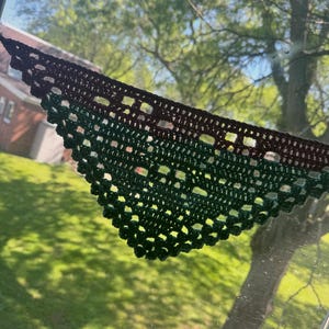 May include: A handmade crochet shawl in a triangular shape. The shawl features a gradient of colors, transitioning from a dark brown at the top to a deep green at the bottom. The open weave design allows light to pass through, creating a delicate, airy appearance.