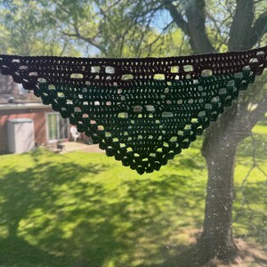 May include: A handmade crochet shawl in a triangular shape, displayed against a window. The shawl transitions from a dark brown top to a gradient of green hues. The open weave design and the ties at the ends are visible.