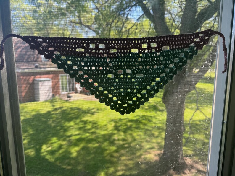 May include: A handmade crochet shawl in a triangular shape, displayed against a window. The shawl transitions from a dark brown top to a gradient of green hues. The open weave design and the ties at the ends are visible.
