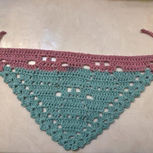 May include: A handmade crochet bandana in a triangular shape. The top section is a dusty rose color, and the bottom is a teal blue. The bandana has ties on each end for securing around the neck.