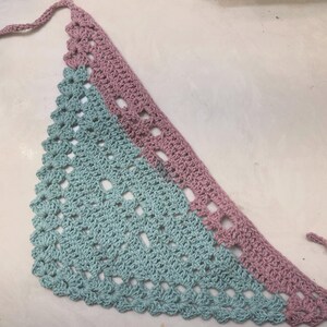 May include: A handmade crochet bandana in a triangular shape. The bandana features a two-tone design, with a light blue and mauve color scheme. The edges are scalloped, and it has ties for securing around the neck.