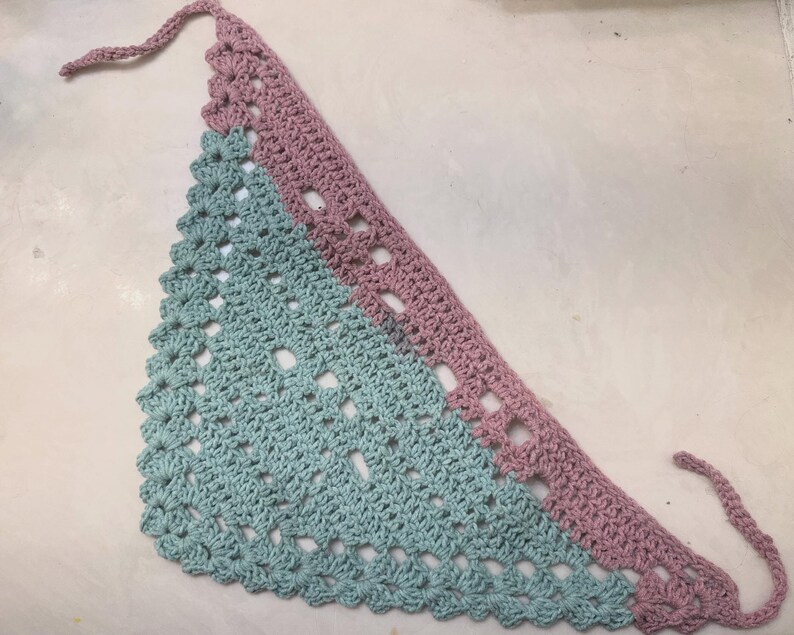 May include: A handmade crochet bandana in a triangular shape. The bandana features a two-tone design, with a light blue and mauve color scheme. The edges are scalloped, and it has ties for securing around the neck.