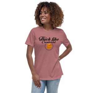 May include: A dusty rose t-shirt with the phrase "Thick Like Cornbread" in black script. The text is above a graphic of a cornbread loaf inside a decorative circle. The shirt has short sleeves and a relaxed fit.
