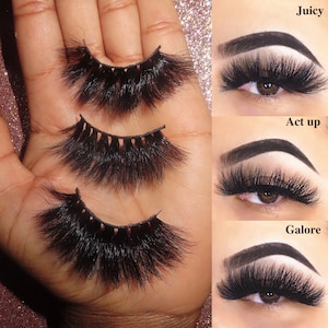Op de afbeelding: Drie paar valse wimpers op een hand, met de labels "Juicy", "Act up" en "Galore".