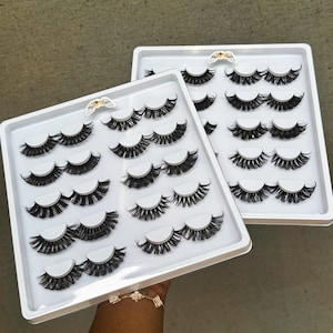 Op de afbeelding: Twee witte kunststof bakjes gevuld met individuele valse wimpers. Elk bakje bevat 10 paar wimpers in verschillende stijlen en lengtes.