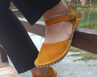 May include: Mustard yellow suede sandals with ankle straps and buckle closures. The sandals have a stitched sole and are worn with black pants. The shoes are on a wooden bridge overlooking a body of water.