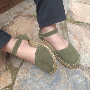 Op de afbeelding: Olijfgroene suède sandalen met enkelbandjes. De sandalen hebben een ronde neus en een gestikte zool. De bandjes zijn vastgezet met een kleine gesp. De schoenen worden gedragen met een zwarte broek.