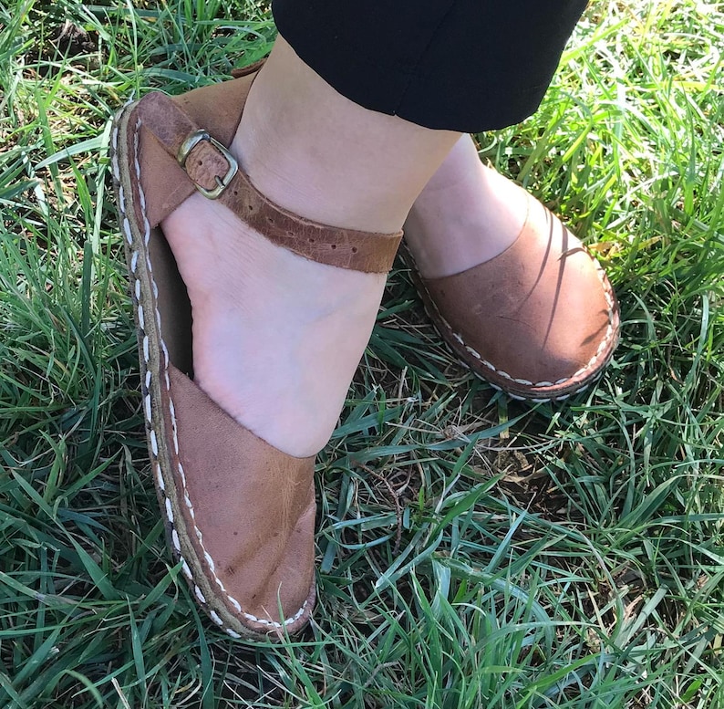 May include: A pair of brown leather sandals featuring a rounded toe, an ankle strap with a metallic buckle, and visible white stitching along the sole edge. The sandals have a flat construction and a smooth, matte finish on the leather surface.