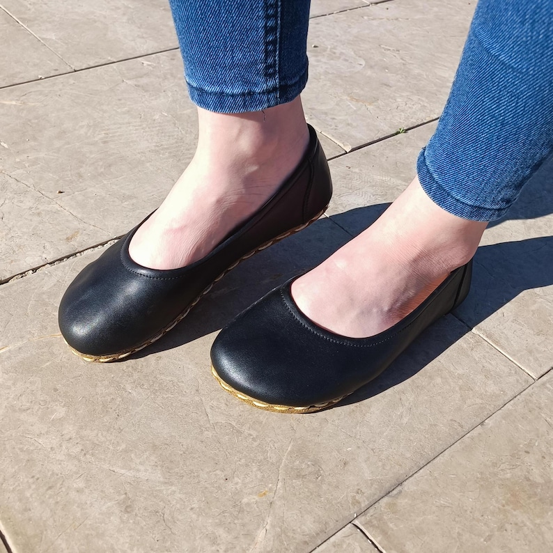 May include: A pair of black leather ballet flats with gold trim around the edges. The flats are worn by a person wearing blue jeans.