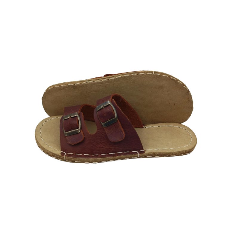 May include: Burgundy leather sandals with two adjustable buckles. The sandals have a light brown sole with white stitching around the edges. The second sandal is upside down, showing the light brown sole.