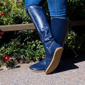 Può includere: Stivali alti blu navy con cerniera laterale. Gli stivali sono realizzati in materiale liscio e hanno una suola marrone chiaro. Gli stivali sono indossati con jeans blu. Gli stivali sono alti circa 40 cm.