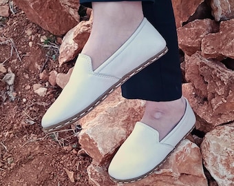 May include: A pair of white leather slip-on shoes with a brown stitch detail around the sole. The shoes are worn on a rocky surface.