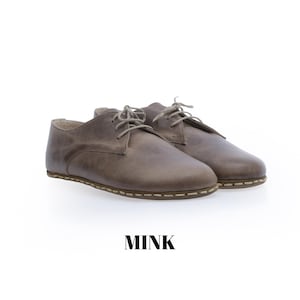 May include: A pair of low-top lace-up shoes in a muted mink brown colour. The shoes feature a smooth leather-like upper, rounded toes, and visible light-coloured stitching along the sole edge. The word MINK is printed in black below the shoes.