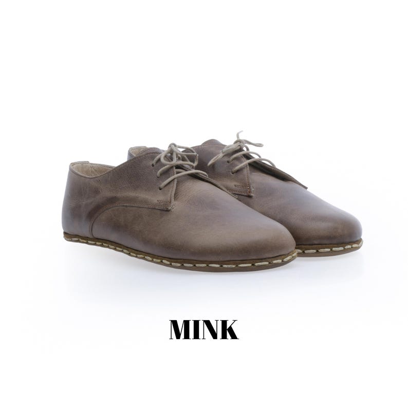 May include: A pair of low-top lace-up shoes in a muted mink brown colour. The shoes feature a smooth leather-like upper, rounded toes, and visible light-coloured stitching along the sole edge. The word MINK is printed in black below the shoes.