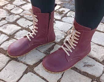 May include: Burgundy leather ankle boots with white laces and a zipper on the side. The boots have a flat sole and are worn on a person's feet.