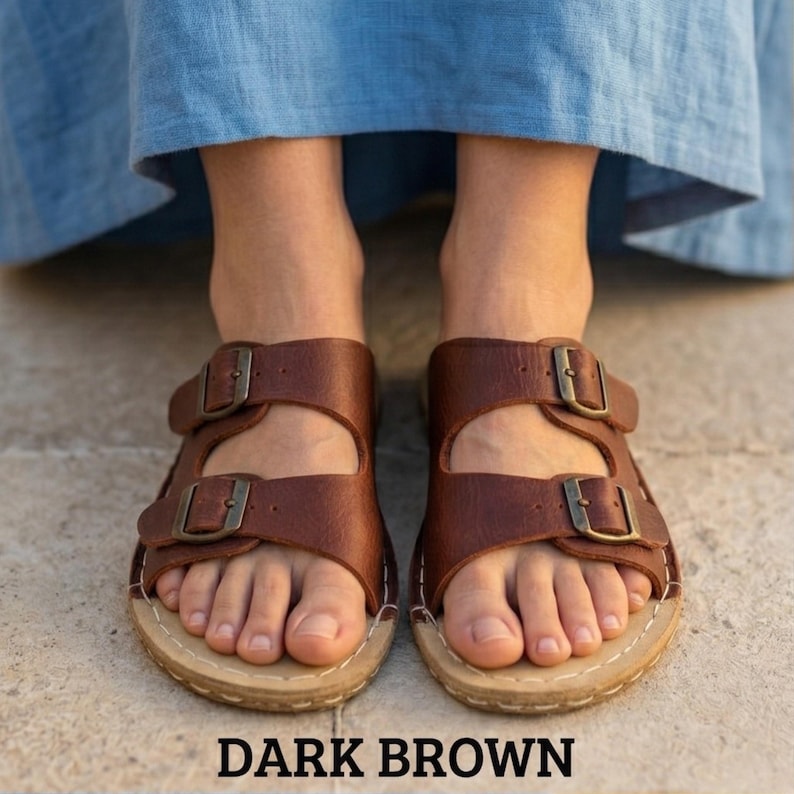 May include: Dark brown leather sandals with two adjustable straps and metal buckles. The sandals have a light-colored sole with visible stitching. The text "DARK BROWN" is displayed below the sandals.