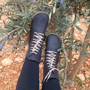 May include: A pair of black leather lace-up boots with beige laces. The boots are worn by a person sitting on the ground near a bush with green leaves and dark blue berries.