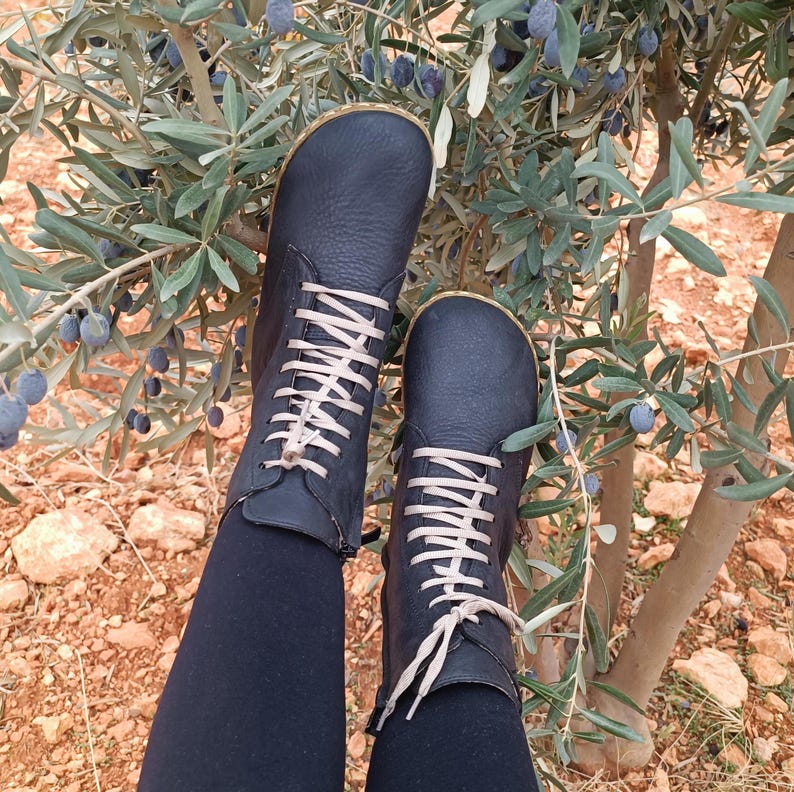 May include: A pair of black leather lace-up boots with beige laces. The boots are worn by a person sitting on the ground near a bush with green leaves and dark blue berries.
