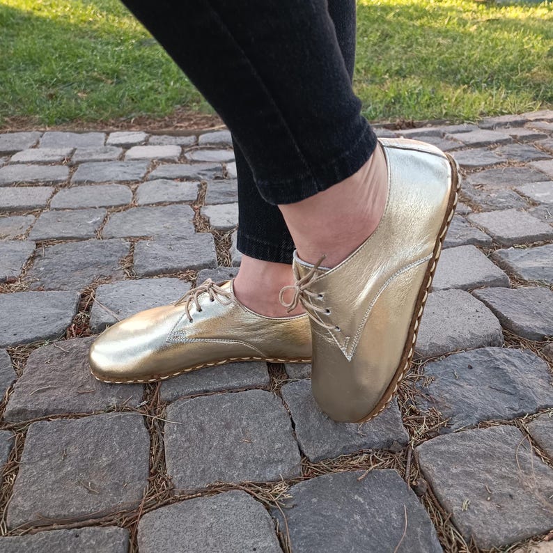 May include: A pair of metallic gold lace-up shoes with a rounded toe and visible stitching along the sole. The shoes feature a low-cut design with thin matching laces and a smooth, reflective finish, worn with dark trousers.