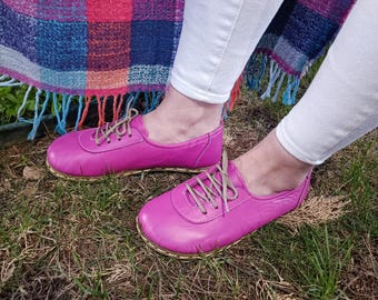 May include: A pair of bright pink leather lace-up shoes with a light brown sole. The shoes are worn by a person sitting on a plaid blanket.