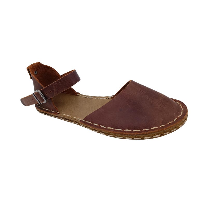 May include: A brown leather sandal featuring a closed rounded toe, an open side design, and a heel strap with a metal buckle. The shoe is constructed with visible light-colored stitching along the perimeter of the sole and a tan inner lining.