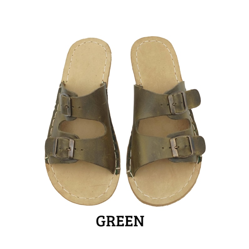 May include: A pair of green leather sandals with two adjustable buckle straps. The sandals have a light brown footbed and white stitching around the edges. The word "GREEN" is printed in black at the bottom.
