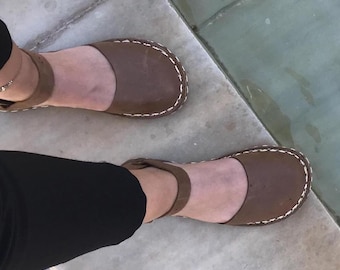 May include: A pair of brown leather flat shoes featuring a rounded toe, an ankle strap with a buckle closure, and visible white contrast stitching along the perimeter of the sole. The shoes have a smooth finish and a low-profile design.