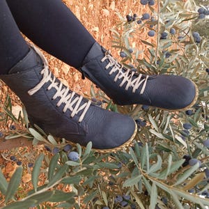 May include: A pair of black leather ankle boots with beige laces. The boots are worn by a person standing on a branch of a tree with green leaves and dark blue berries.
