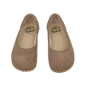 May include: A pair of taupe-colored, suede-like ballet flats. The shoes have a rounded toe and a stitched sole. The interior of each shoe features the text "Galle Barefoot Handmade".