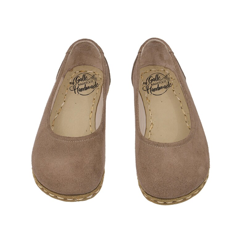 May include: A pair of taupe-colored, suede-like ballet flats. The shoes have a rounded toe and a stitched sole. The interior of each shoe features the text "Galle Barefoot Handmade".
