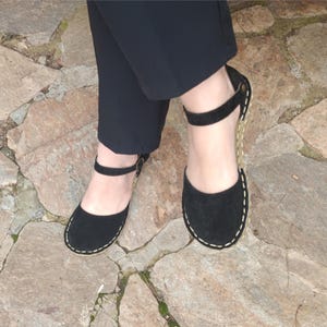 Op de afbeelding: Zwarte suède sandalen met een band en een gespsluiting. De sandalen hebben een jute zool en worden gedragen op een stenen oppervlak.