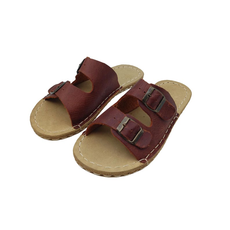 May include: A pair of brown leather sandals with two straps and metal buckles. The sandals have a light tan sole and are stitched with white thread. The footwear is designed for casual wear.
